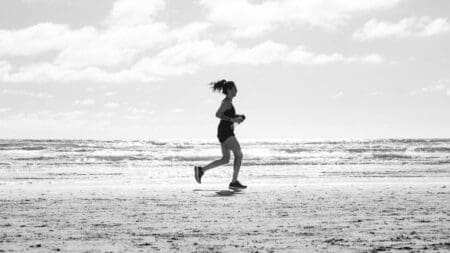 6 Best Running Shoes for Beginners (2026) Black and white photograph of a woman running along the sandy shoreline of an ocean beach