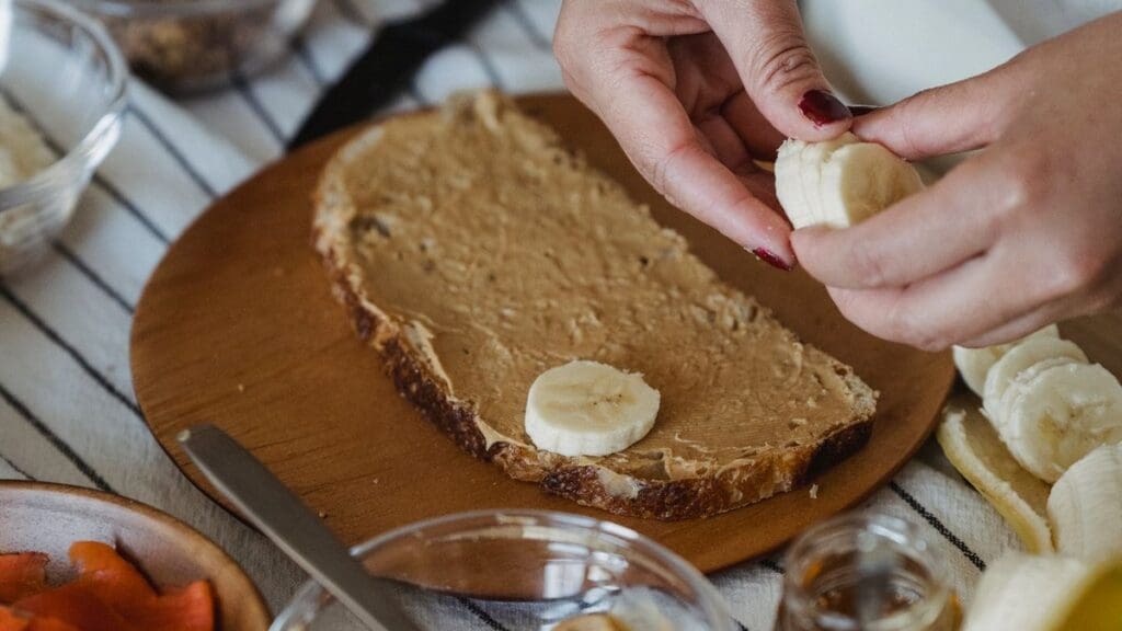 How Long After Eating Should You Run? (+ Pre-Run Planner) Hands placing fresh banana slices onto peanut butter toast on a round wooden board for a pre-run snack