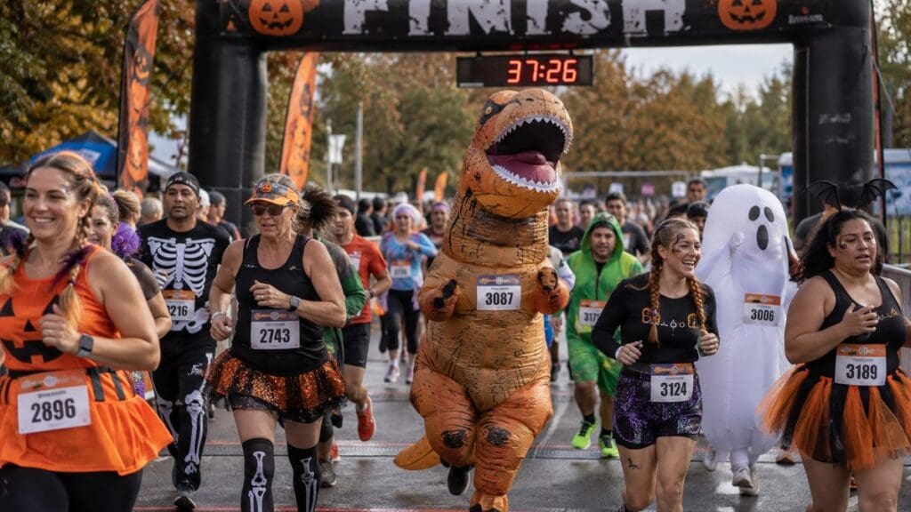 22 Best Inflatable Costumes for a Halloween 5K: Funny, Viral & Completely Unhinged Runner in an inflatable T-Rex costume and runners in Halloween outfits crossing a 5K race finish line arch