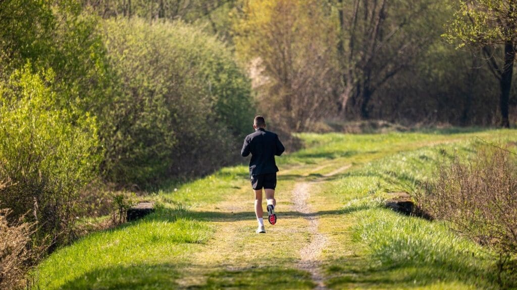 25 Running Goals That Have Nothing to Do With Getting Faster Rear view of a runner jogging down a tranquil nature trail surrounded by lush green trees and warm morning light