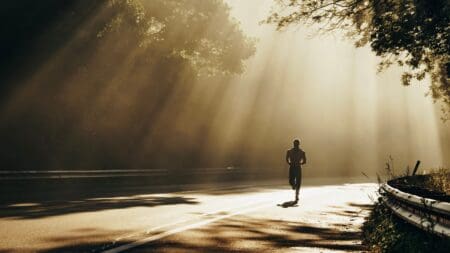 Running With Plantar Fasciitis: Manage Heel Pain & Return to Running Runner in athletic gear jogs on a paved forest road, with warm morning sunbeams streaming through tall trees.